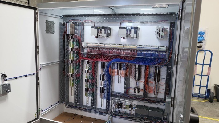 An open control panel wired to a drawing, with PLC racks and terminal strips